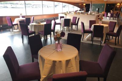 Terrasse d'un bateau avec des fauteuils, des plantes décoratives et une vue sur un pont et des arbres en arrière-plan.