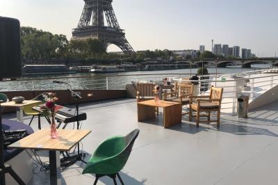 Terrasse d'un bateau avec des fauteuils, des plantes décoratives et une vue sur un pont et des arbres en arrière-plan.