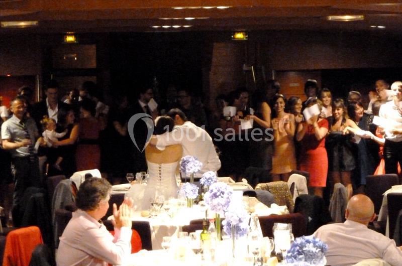 Un couple de mariés s'embrasse au centre d'une salle de réception, entouré d'invités applaudissant.