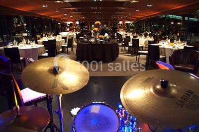 Terrasse d'un bateau avec des fauteuils, des plantes décoratives et une vue sur un pont et des arbres en arrière-plan.