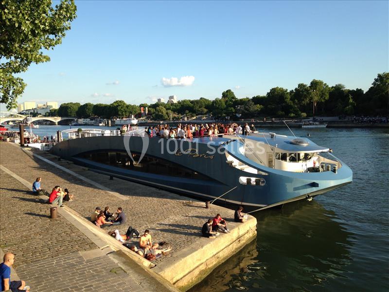 Bateau moderne amarré sur la Seine avec des passagers à bord et des personnes assises sur les quais ensoleillés.