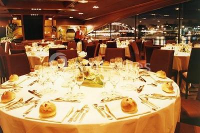 Terrasse d'un bateau avec des fauteuils, des plantes décoratives et une vue sur un pont et des arbres en arrière-plan.