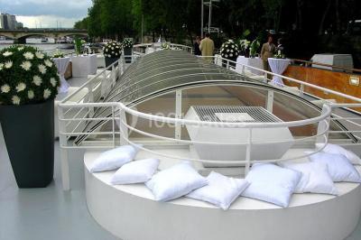 Terrasse d'un bateau avec des fauteuils, des plantes décoratives et une vue sur un pont et des arbres en arrière-plan.