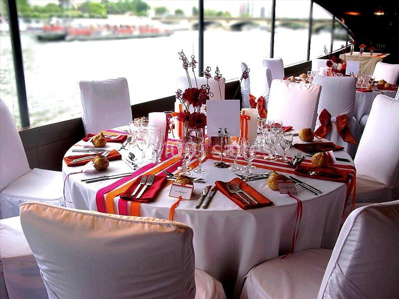 Table ronde élégamment dressée avec nappes blanches, rubans colorés, vaisselle raffinée et vue sur un fleuve.