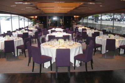 Terrasse d'un bateau avec des fauteuils, des plantes décoratives et une vue sur un pont et des arbres en arrière-plan.