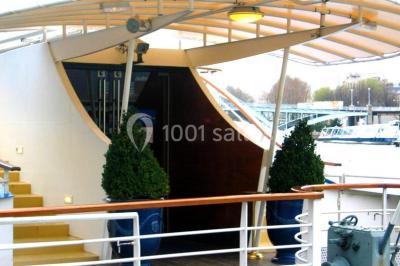 Terrasse d'un bateau avec des fauteuils, des plantes décoratives et une vue sur un pont et des arbres en arrière-plan.