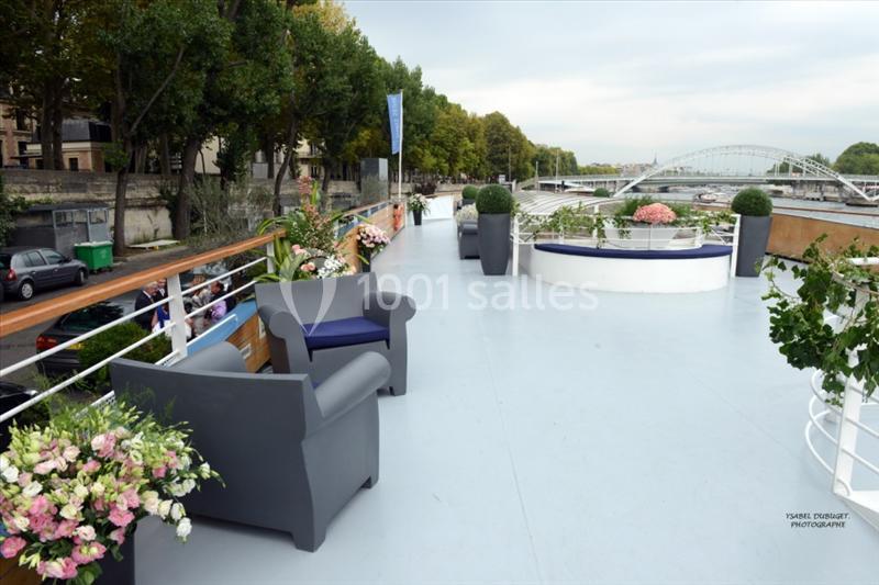 Pont supérieur d'un bateau avec fauteuils, plantes décoratives et vue sur un fleuve bordé d'arbres et de ponts.