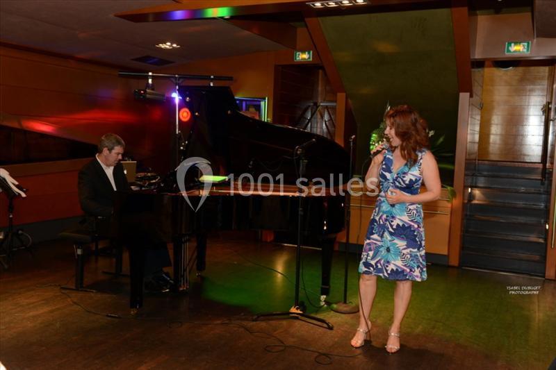 Une femme chante sur scène près d'un pianiste dans une salle éclairée aux tons chauds.