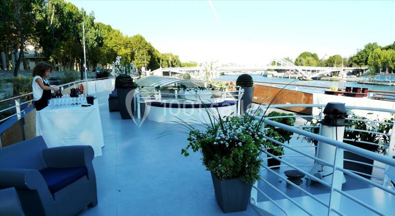 Terrasse d'un bateau avec des plantes, des fauteuils et une table dressée, vue sur la Seine et des ponts en arrière-plan.