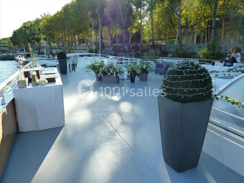 Terrasse d'un bateau aménagée avec des plantes, des tables et des décorations, bordée par des arbres au bord de l'eau.