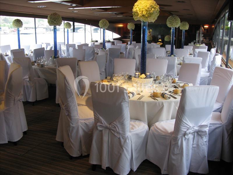 Salle de réception décorée avec des tables rondes, nappes blanches, chaises houssées et centres de table fleuris.