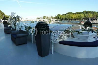 Terrasse d'un bateau avec des fauteuils, des plantes décoratives et une vue sur un pont et des arbres en arrière-plan.