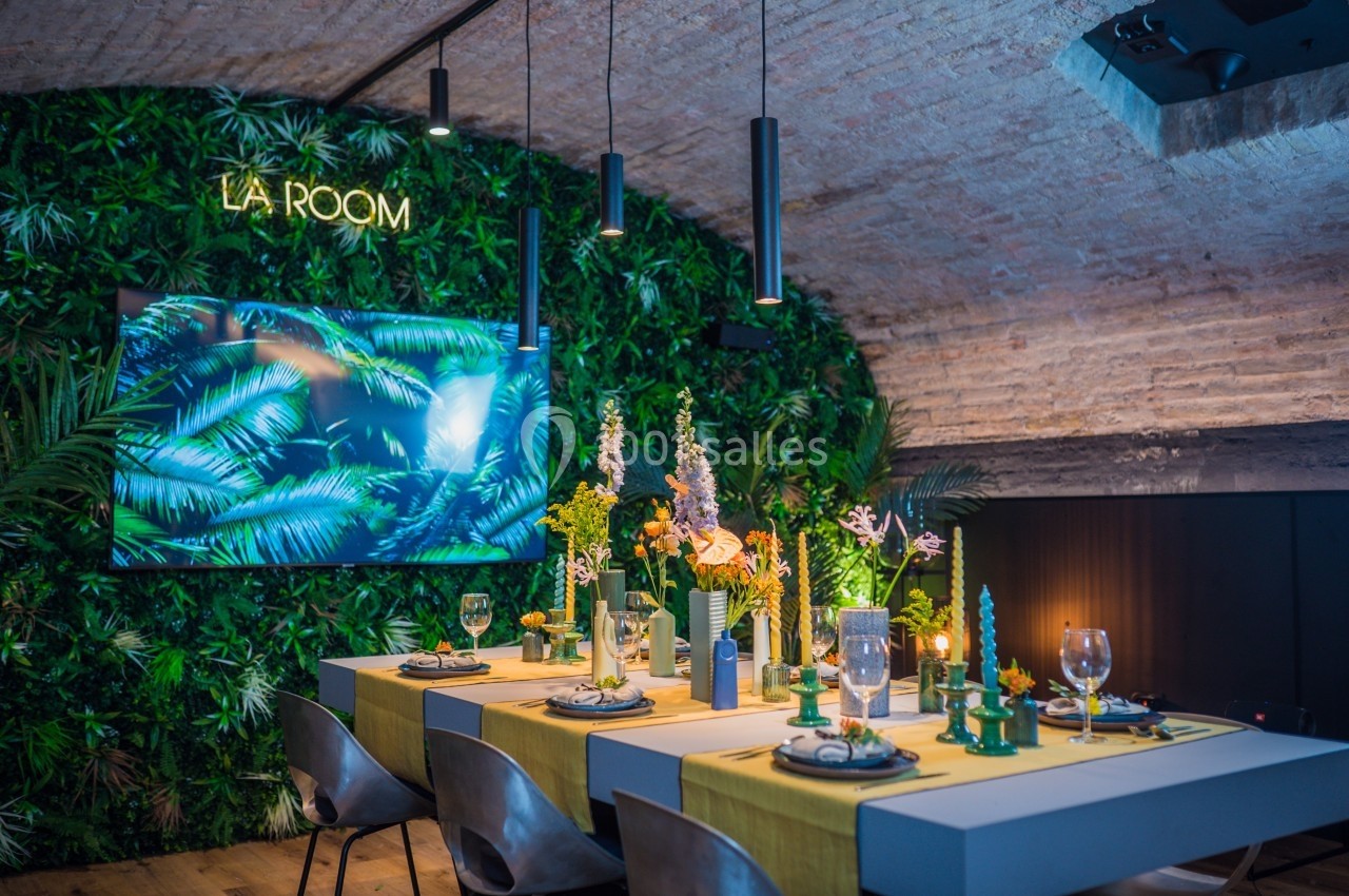 Salle voûtée décorée de plantes, avec une table dressée pour un repas et un écran affichant des feuilles tropicales.