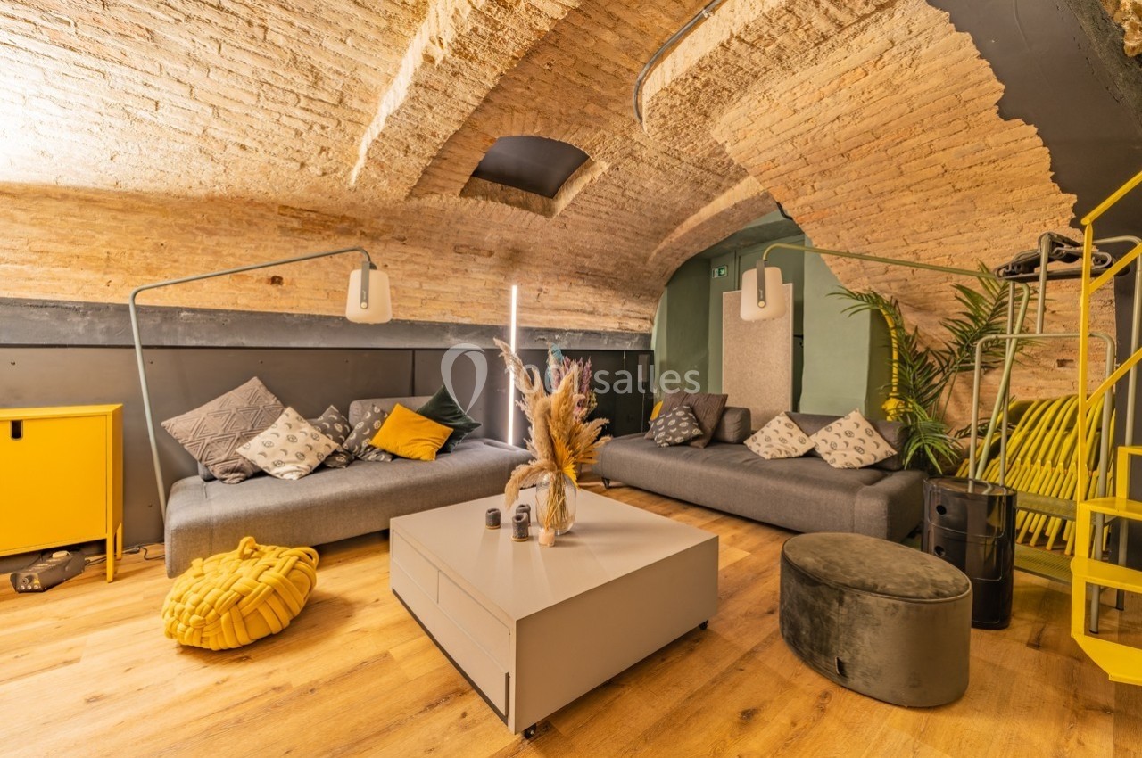 Salon voûté avec murs en briques, canapés gris, table basse moderne et décoration chaleureuse dans des tons jaunes et gris.