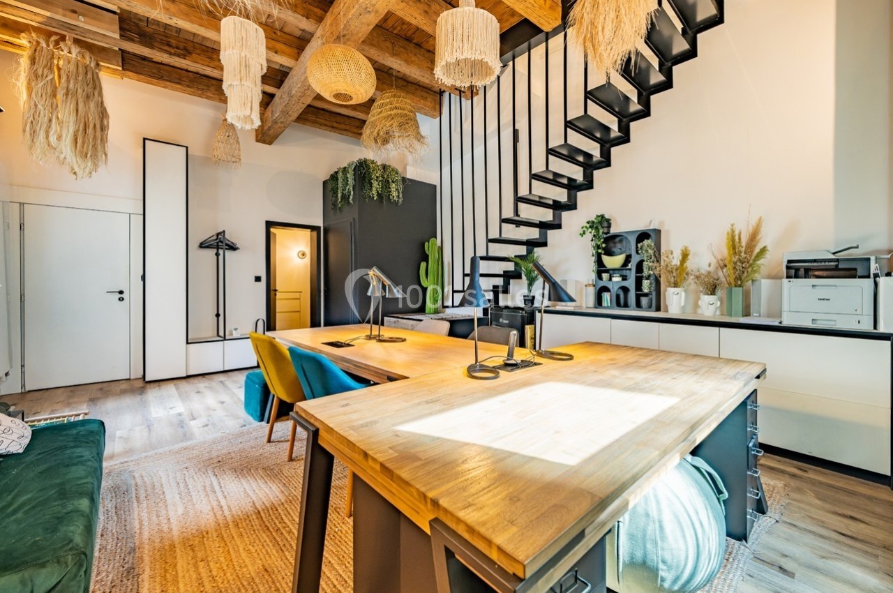 Espace de travail lumineux avec table en bois, chaises colorées, escalier métallique et décoration naturelle suspendue.