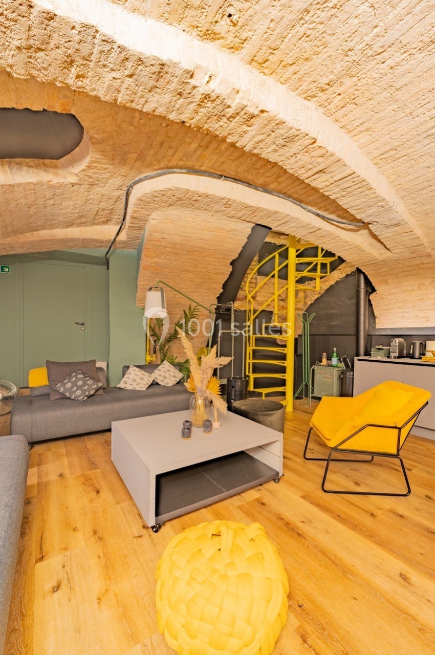 Salon voûté avec murs en pierre, mobilier moderne jaune et gris, escalier métallique et parquet en bois clair.
