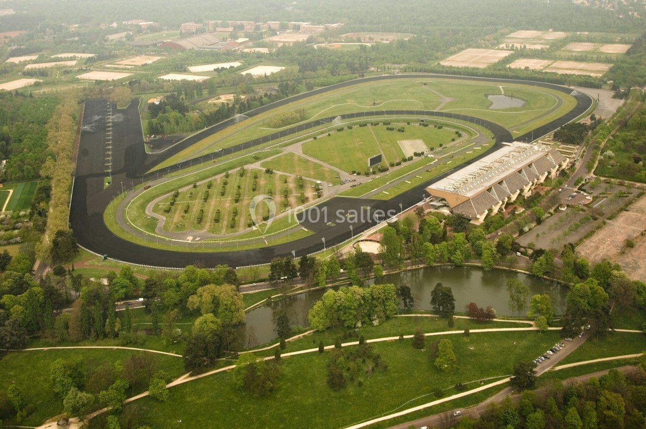 Vue aérienne d'un circuit automobile entouré de verdure, avec des bâtiments et un plan d'eau à proximité.
