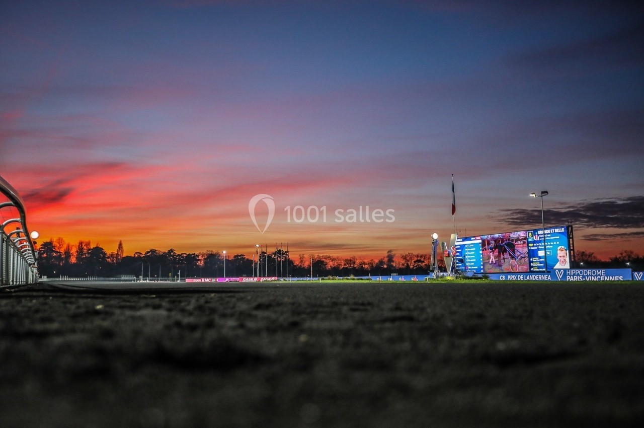 Coucher de soleil vibrant sur un hippodrome, avec écran lumineux et piste en premier plan.