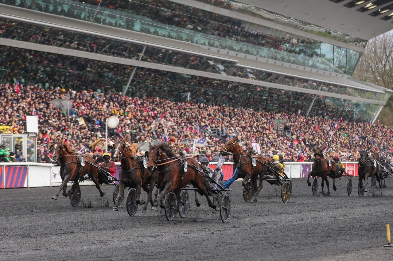Course de trot attelé sur une piste devant des tribunes pleines de spectateurs.