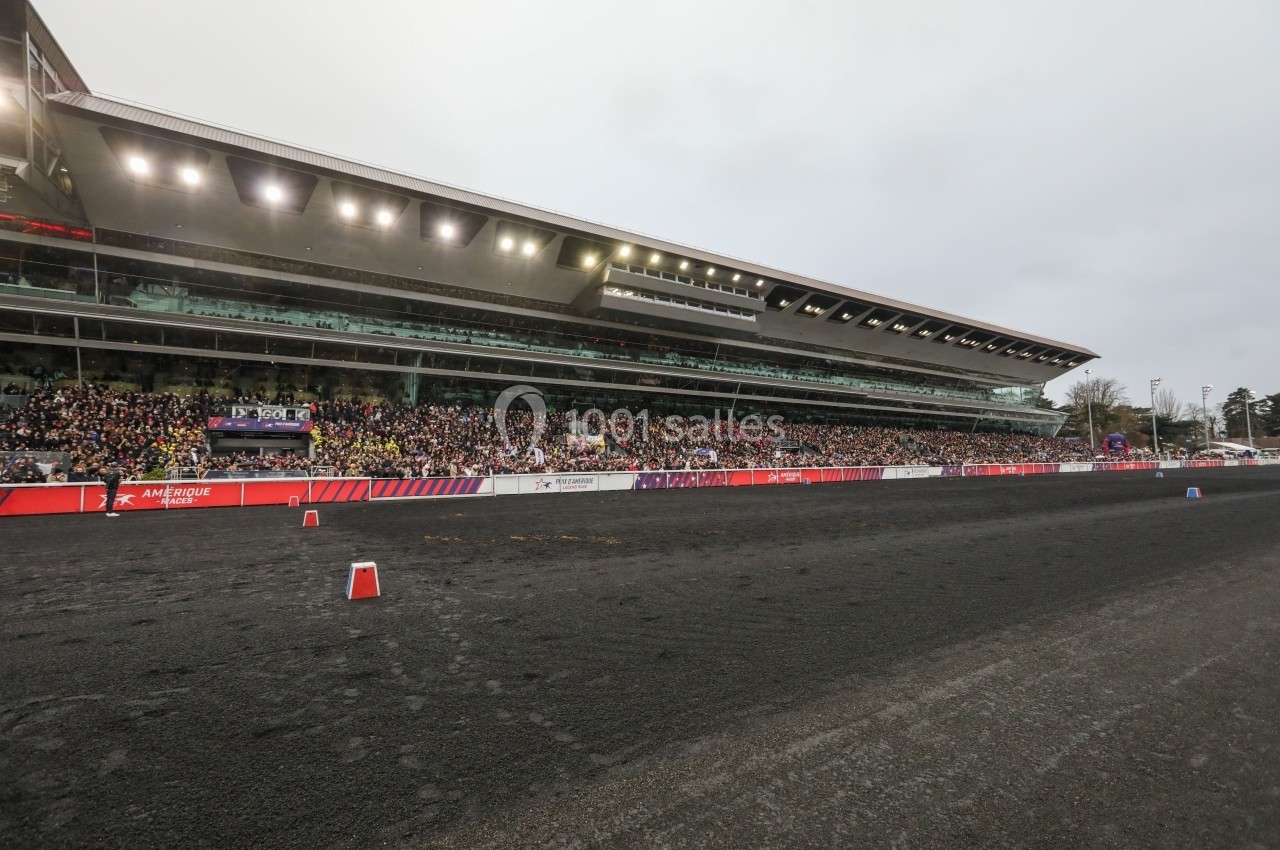 Tribune bondée d'un hippodrome moderne avec une piste sombre au premier plan, sous un ciel nuageux.