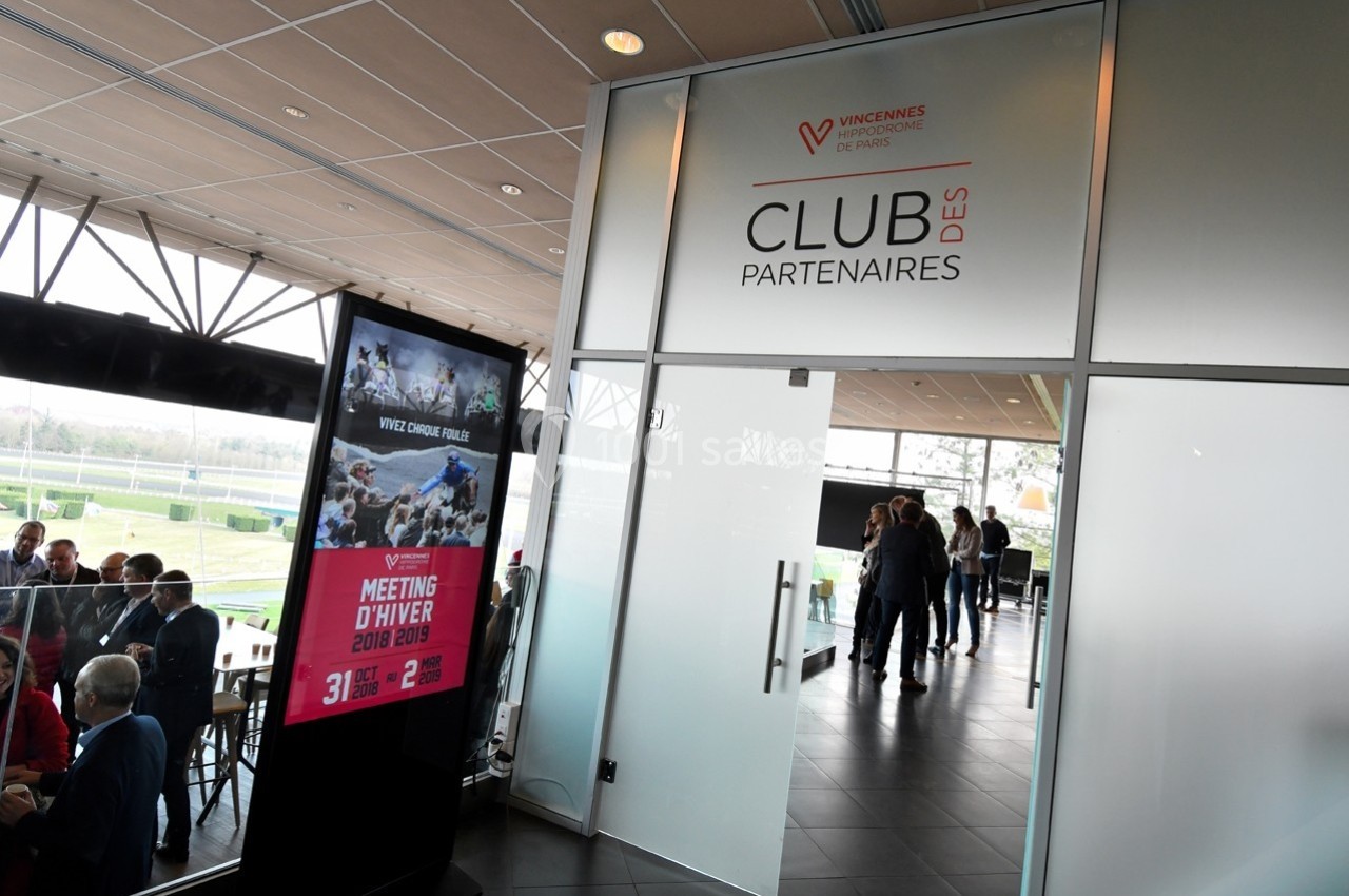 Entrée vitrée du Club des Partenaires avec des affiches d'événements et des personnes discutant à l'intérieur.