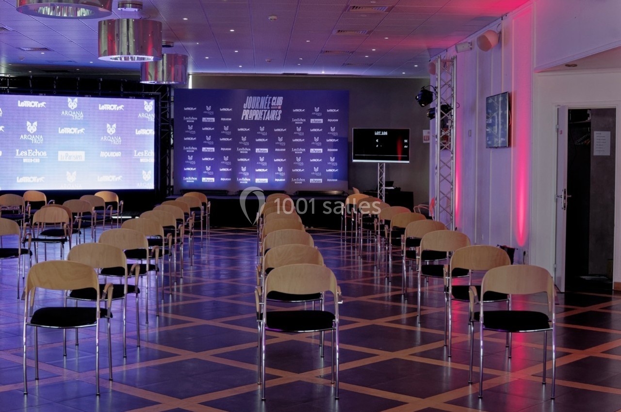 Salle de conférence avec des rangées de chaises alignées face à une scène équipée d'écrans et d'un fond personnalisé.