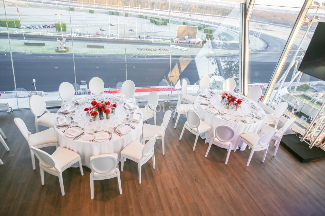 Tables rondes élégamment dressées avec nappes blanches et bouquets de fleurs rouges, vue sur un hippodrome en arrière-plan.