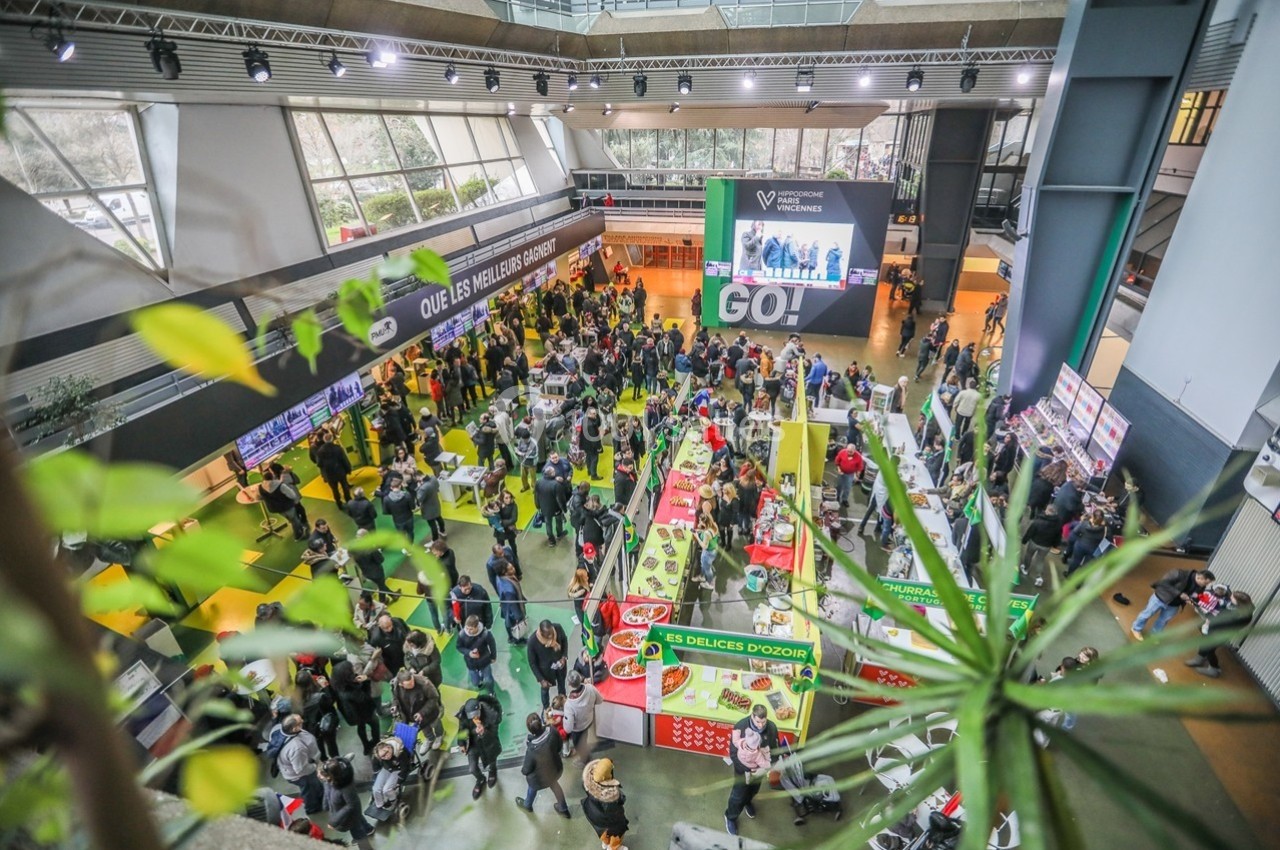 Vue aérienne d'un salon animé avec des stands colorés, des visiteurs et une grande bannière affichant ’GO!’.
