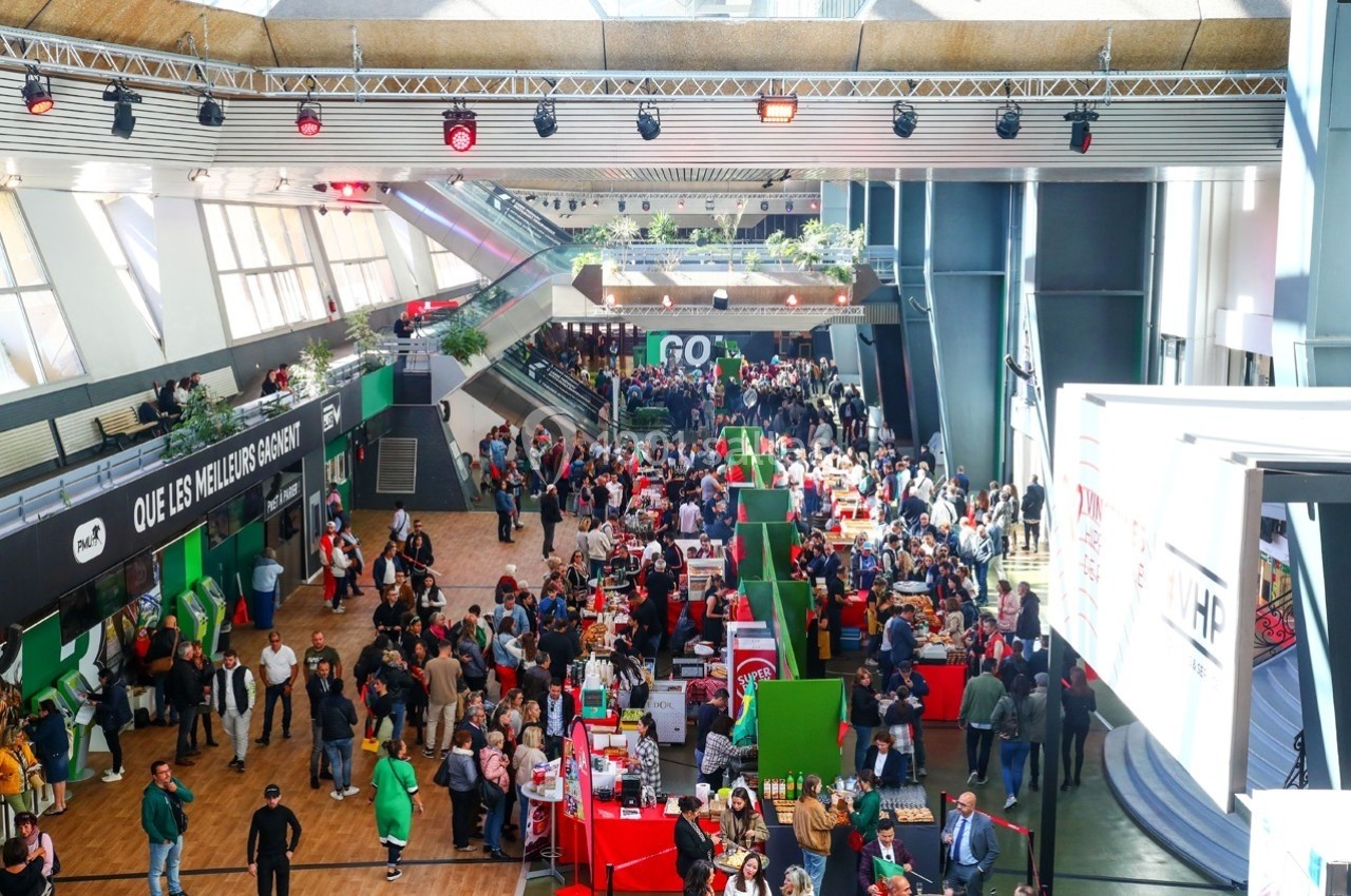 Foule visitant un salon avec stands colorés, éclairage naturel et mezzanine en arrière-plan.