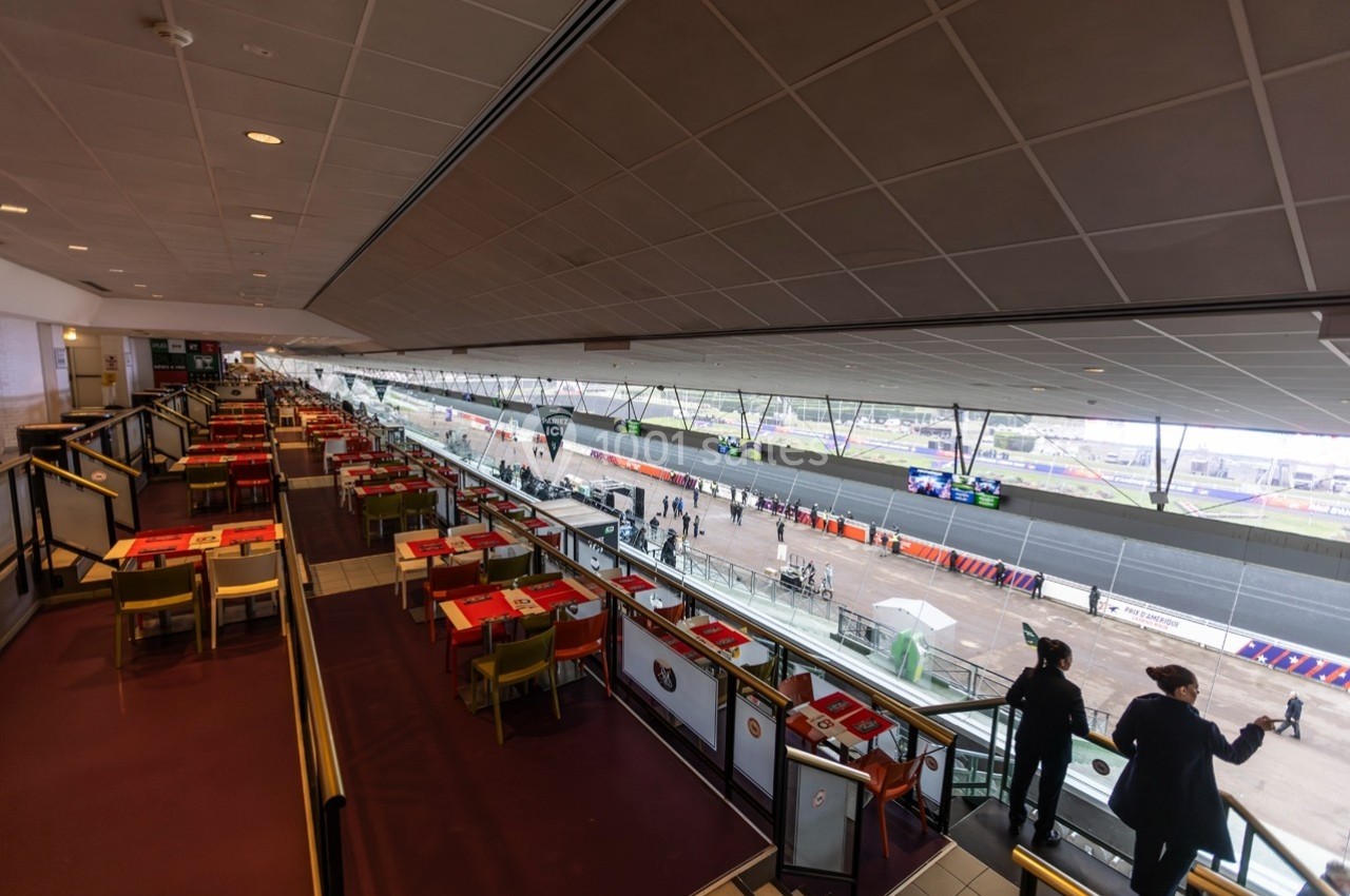 Salle de restaurant avec vue panoramique sur une piste de course, tables dressées et quelques personnes en contrebas.
