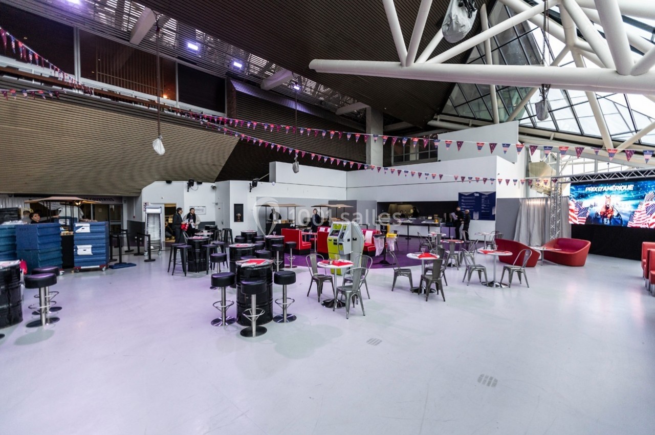 Salle spacieuse avec tables, chaises et décorations suspendues, éclairée par des lumières artificielles et naturelles.
