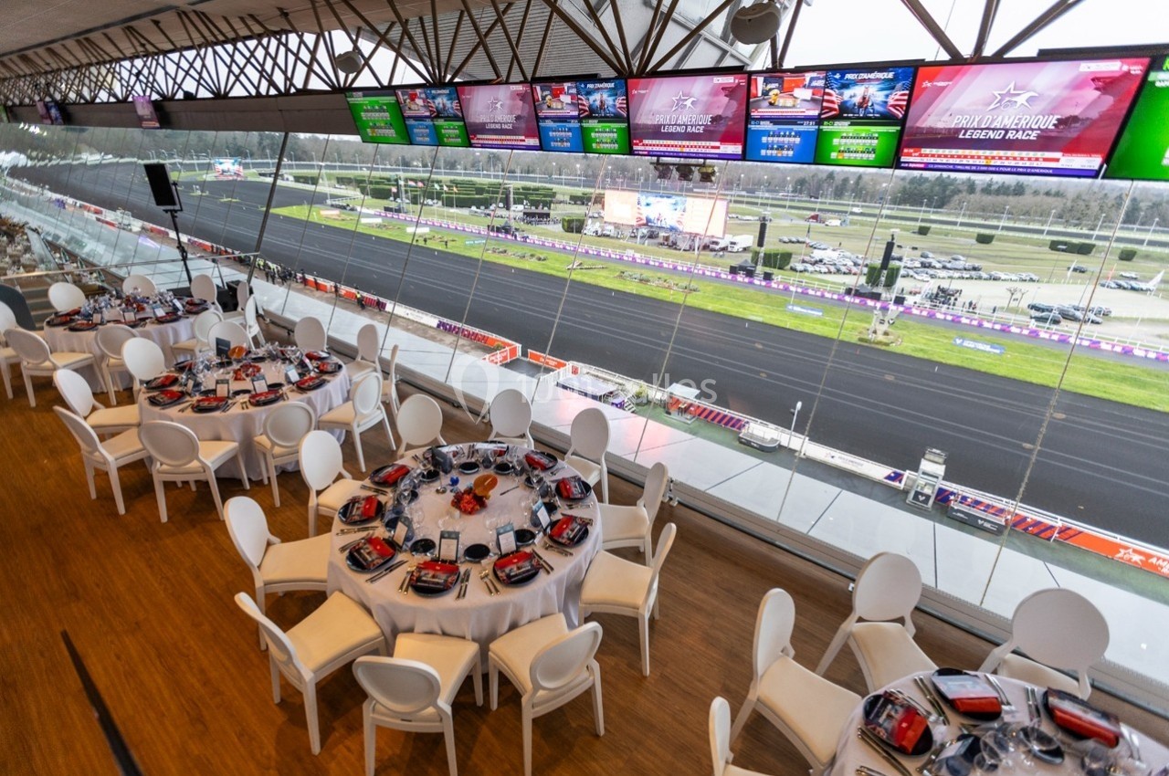 Salle de réception avec tables dressées, vue panoramique sur une piste de course et écrans diffusant des images.