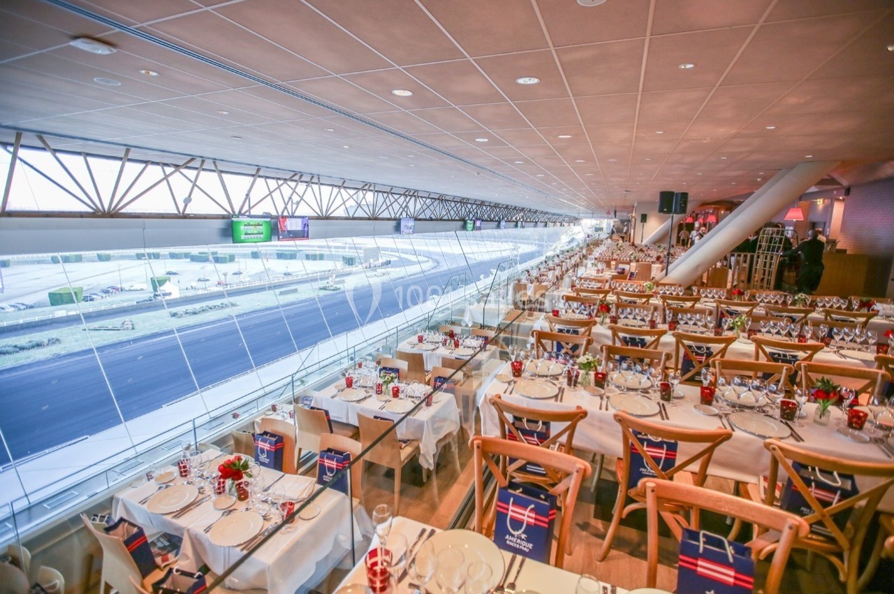 Salle de restaurant lumineuse avec vue sur un circuit, tables dressées avec nappes blanches et décorations.