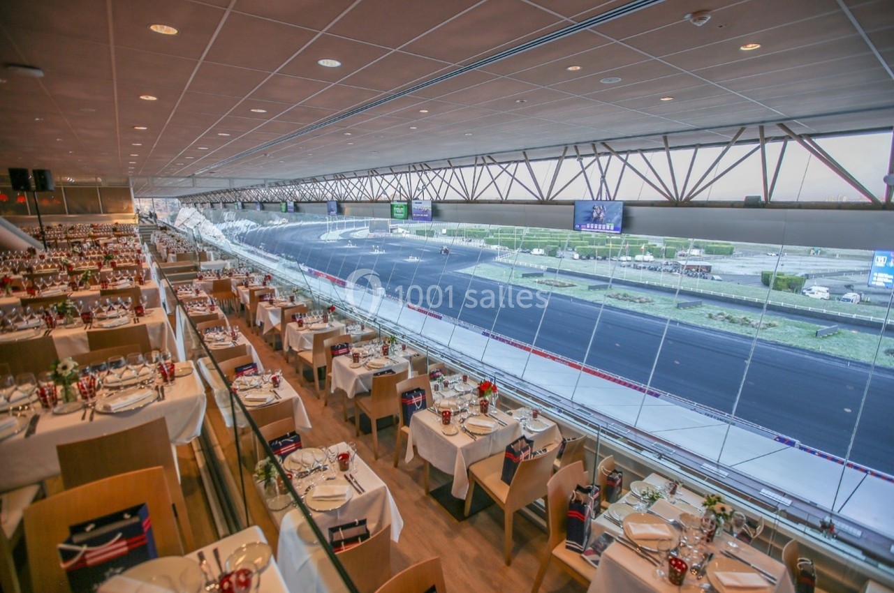 Salle de restaurant avec vue panoramique sur un circuit automobile, tables dressées et baies vitrées.