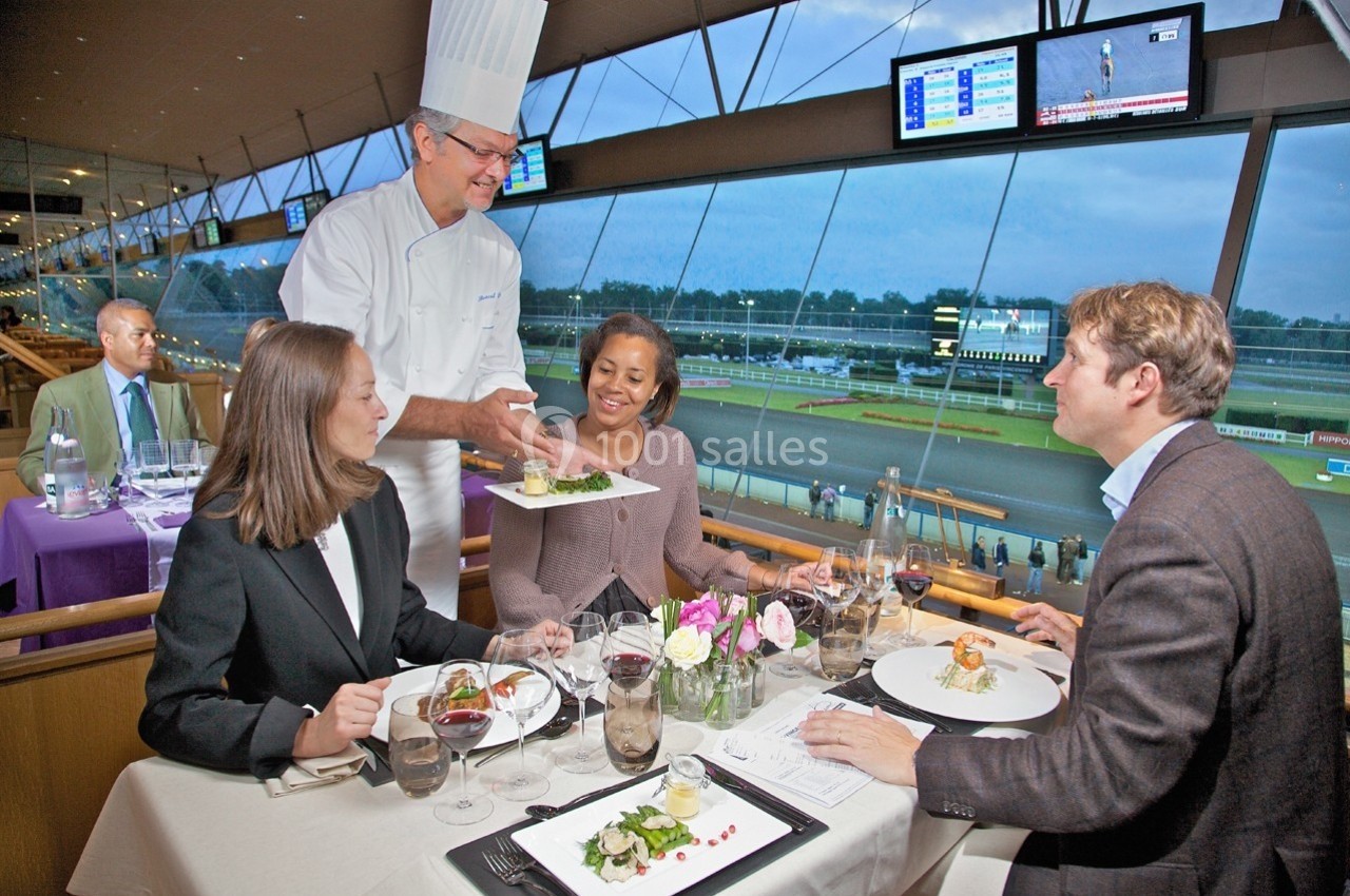Un chef sert des plats à trois convives attablés dans un restaurant avec vue sur un hippodrome.