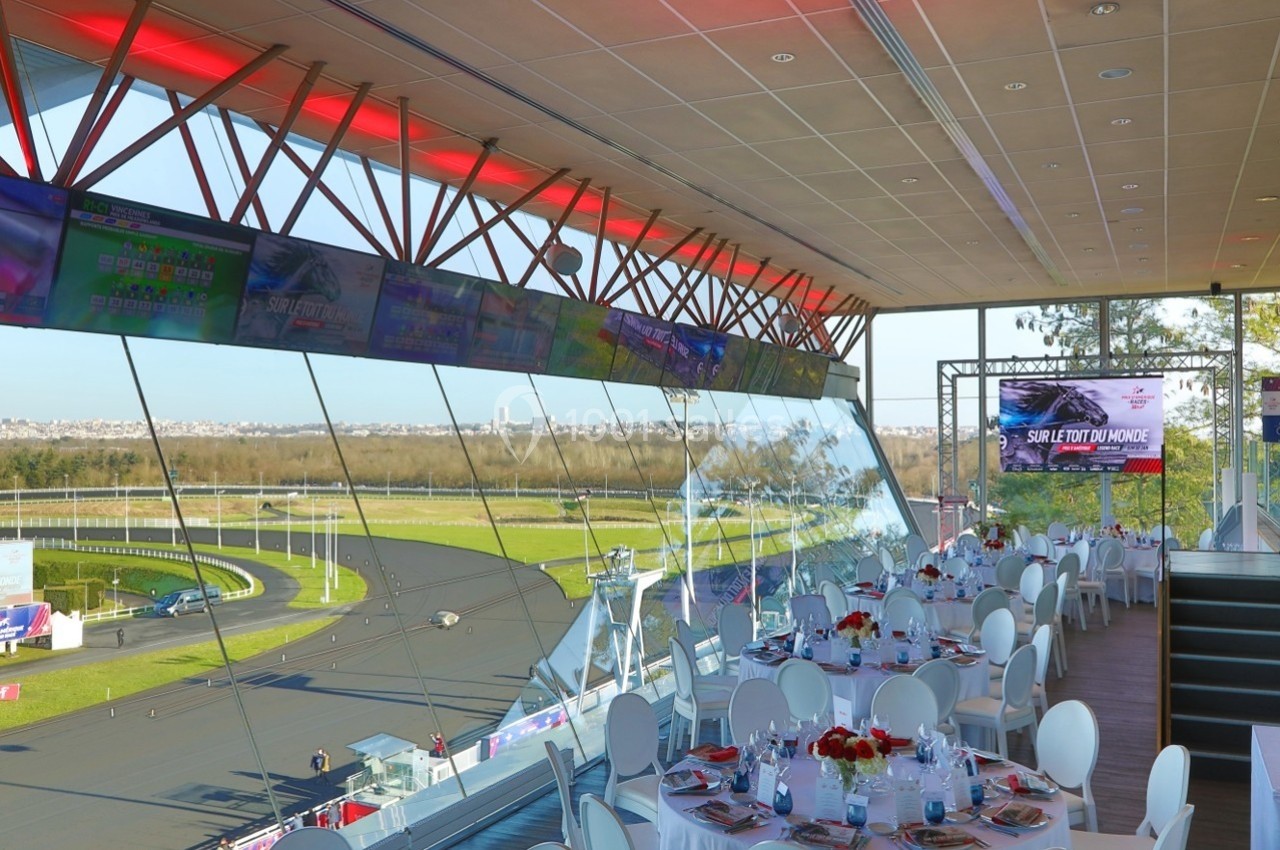 Salle de réception avec tables dressées, vue panoramique sur un circuit de course et écrans suspendus diffusant des images.