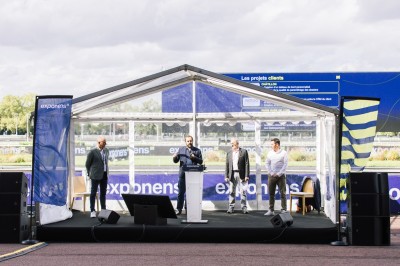 Trois personnes sur une scène extérieure sous une tente, devant un écran affichant des données financières.