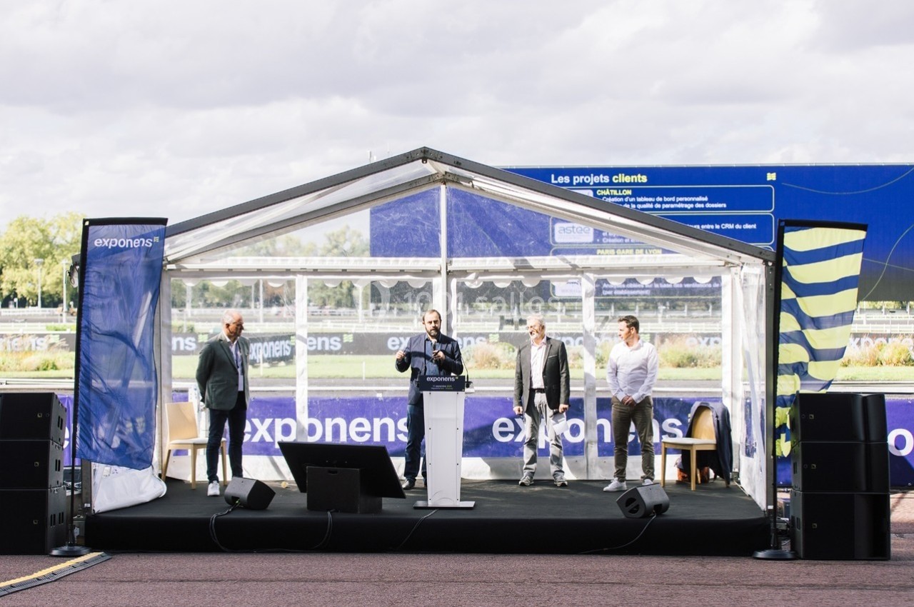 Quatre hommes sur une scène sous une tente transparente, avec un écran et des bannières en arrière-plan.