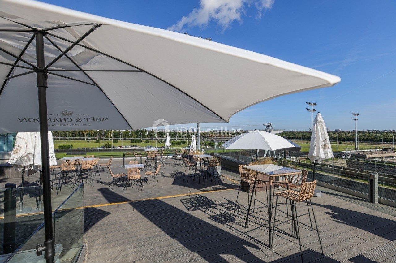 Terrasse ensoleillée avec tables, chaises en rotin et parasols blancs, offrant une vue dégagée sur un paysage verdoyant.