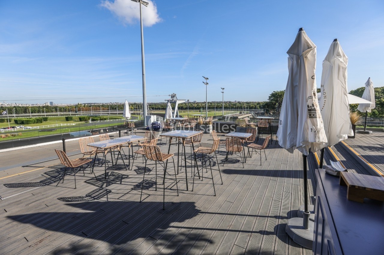 Terrasse extérieure avec tables et chaises en métal, parasols fermés, vue sur un espace vert et ciel dégagé.