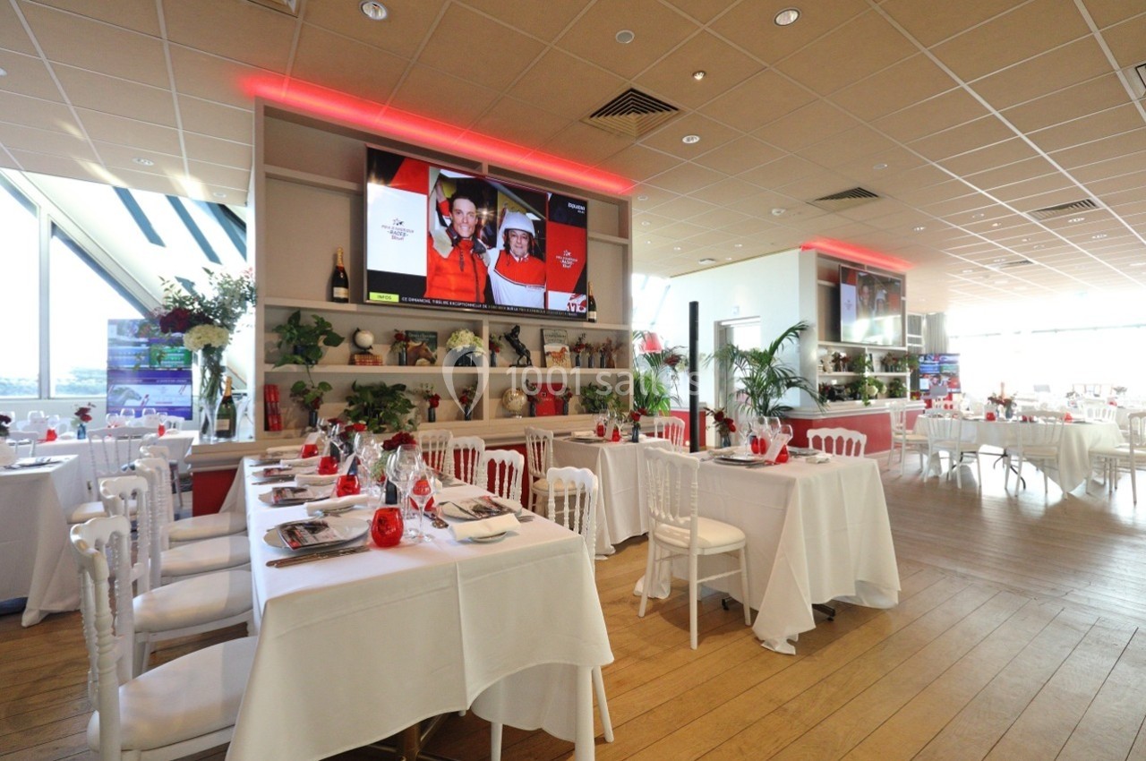 Salle de restaurant lumineuse avec tables dressées, chaises blanches et écrans diffusant des images en arrière-plan.