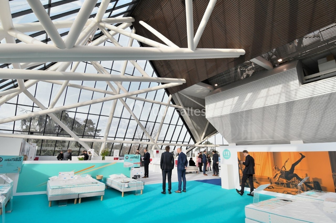 Vue d'un espace d'exposition lumineux avec des lits exposés, des visiteurs discutant et une structure moderne en arrière…