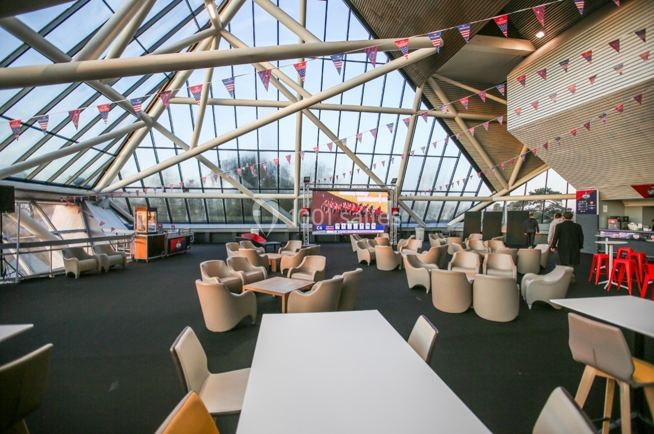 Salle lumineuse avec de grandes baies vitrées, des fauteuils et des tables, décorée de fanions suspendus.