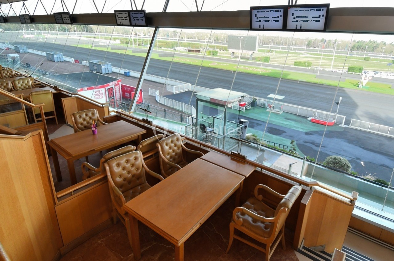 Salle avec des tables et fauteuils en cuir, offrant une vue panoramique sur une piste de course à travers de grandes baies…