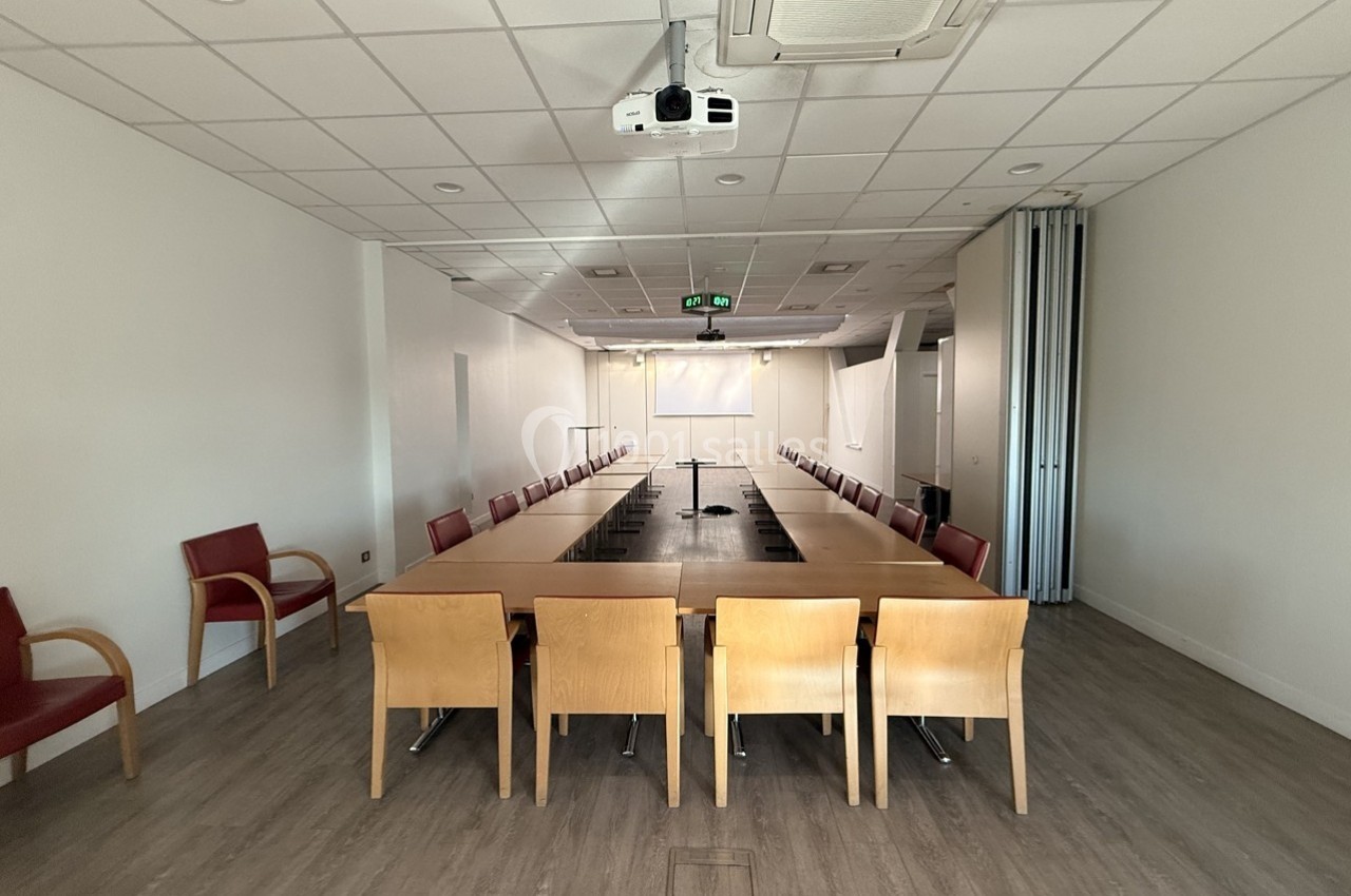 Salle de réunion avec une grande table rectangulaire entourée de chaises, écran de projection et projecteur au plafond.