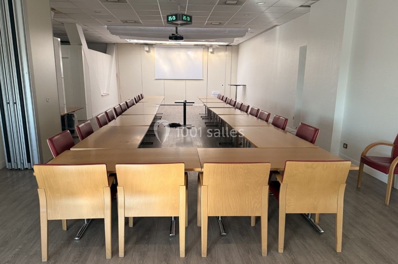 Salle de réunion lumineuse avec tables disposées en U, chaises en bois et écran de projection au fond.
