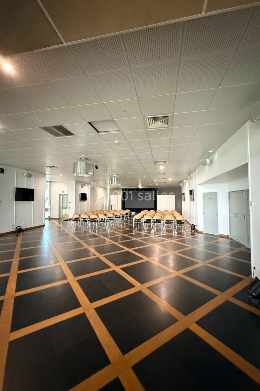 Salle de conférence vide avec des chaises alignées devant un écran, sol en parquet sombre et plafond avec éclairage intégré.
