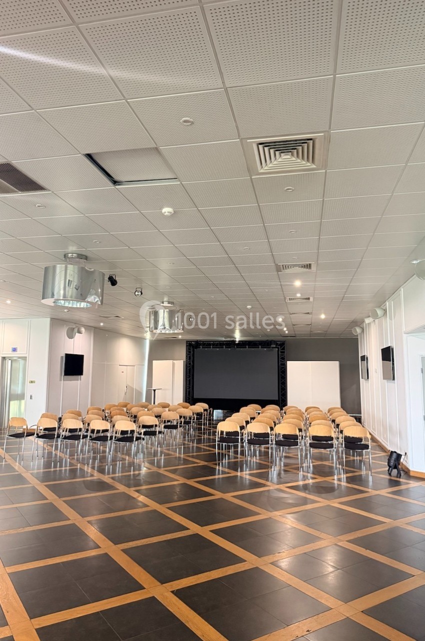 Salle de conférence vide avec des rangées de chaises face à un écran noir, éclairée par des lumières au plafond.