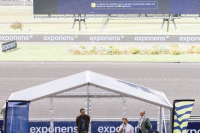 Trois personnes sur une scène extérieure sous une tente, devant un écran affichant des données financières.