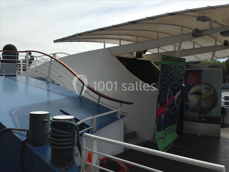 Pont supérieur d'un bateau avec une rampe en bois, des affiches publicitaires et une vue partielle de l'extérieur.